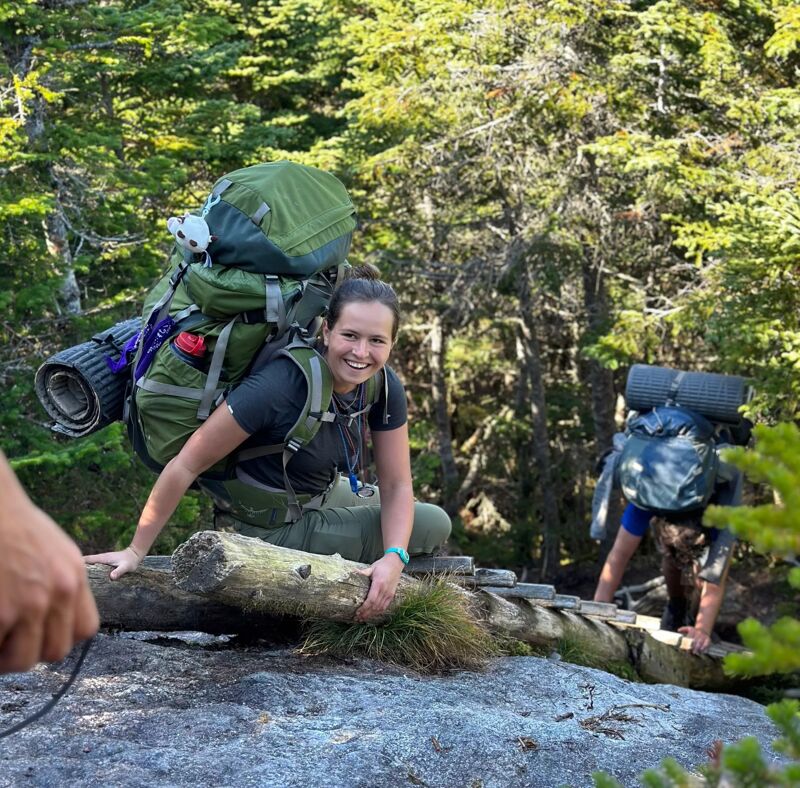 The image shows two people hiking in a forest. The person in the foreground is a woman with a large backpack, smiling at the camera. She is standing on a rocky outcrop with a log bridge. Another person with a backpack is climbing the log bridge behind her. The background is filled with trees and foliage.